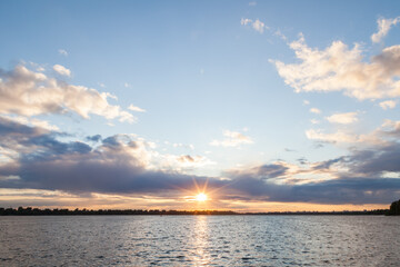Evening landscape with the setting sun on a background of a painted sky in blue, pink and purple colors around a yellow sun over a river or lake.