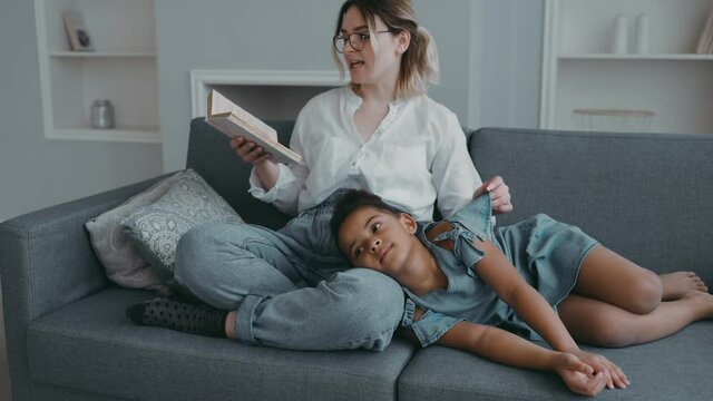 Young caring mother or nanny with glasses, sits on couch in living room with book in her hands and reads her african american girl fascinating fairy tale. Little one listens carefully lying on sofa.