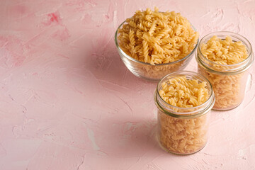 Raw wheat golden spiral pasta in glass bowl and jar on textured pink background, angle view copy space