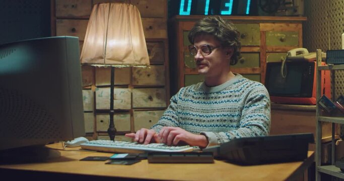 Caucasian male nerd in glasses with mustache sitting at desk in retro room and working on computer. Man programist typing on keyboard, studying and smiling. Vintage style of 80's. Gamer from 90's.