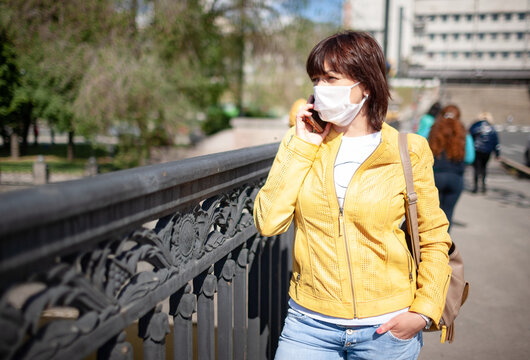 Funny middle-aged woman in a white protective mask talking on a smartphone while walking around the city on a warm spring day. Coronovirus pandemic and remote communication concept