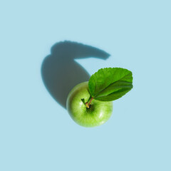 Green apple on stem with leaf on light blue background in bright sunlight with drop shadow. Conceptual minimalist style creative food poster