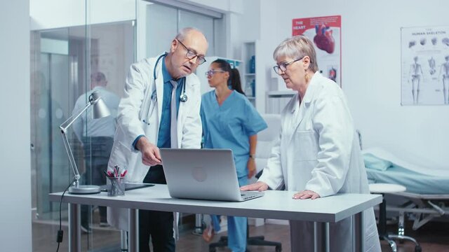 Two Elderly Senior Experienced Doctors Deciding Patient Treatment While Nurse Is Working In Background. Senior Authentic Doctors In Modern Private Hospital Clinic Room Healthcare System Medicine And