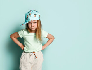 Upset little girl standing with hands on hips, bending brows and looking angrily. Cropped studio shot isolated on light blue, copy space