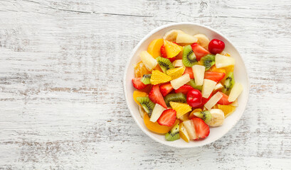 Summer fruit salad in a plate of chopped strawberries, kiwi, banana, orange and cherry on white wooden background. Top view with copy space. Healthy organic food.