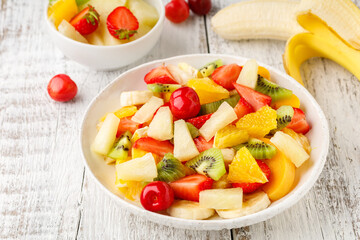 Fruit salad in a plate of chopped strawberries, kiwi, banana, orange and cherry on white wooden background. Healthy organic food.
