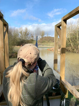 Woman shooting shotgun over water at a clay shooting range. Shot and wadding can be seen flying towards the clay target.
