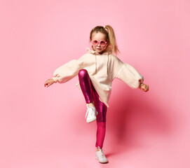 Concentrated little girl posing on one leg raising up her knee and keeping hands wide apart to find balance. Full length shot isolated on pink