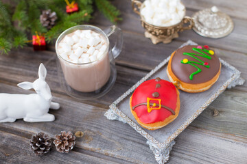Christmas chocolate donuts with Santa Claus icing and new year tree on ceramic stand. Glass cup of cocoa with marshmallows on wooden table. Spruce branch, present, cones, sugar bowl, white deer