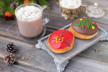 Christmas chocolate donuts with Santa Claus icing and new year tree on ceramic stand. Glass cup of cocoa with marshmallows on wooden table. Spruce branch, present, cones, sugar bowl, white deer