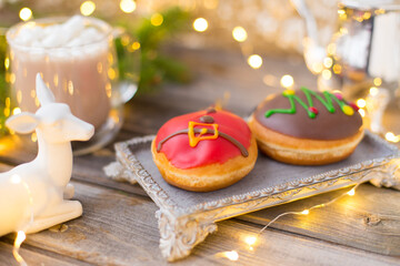 Christmas chocolate donuts with Santa Claus icing and new year tree on ceramic stand. Glass cup of cocoa with marshmallows on wooden table. Spruce branch, present, cone, sugar bowl,white deer,garland