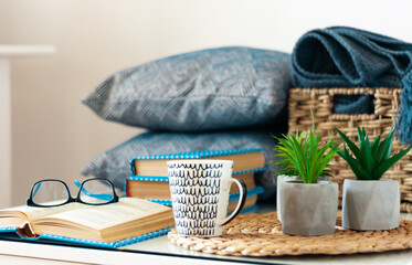 Cozy home interior decor: stack of books, plants in pots on a wicker stand, pillows and plaid on a white table. Distance home education. Quarantine concept of stay home.