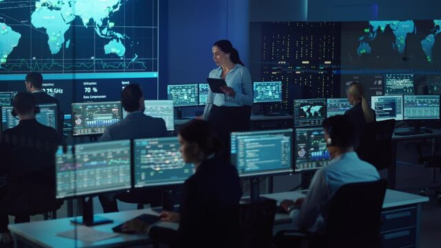 Female Project Leader with Tablet Computer Walks to a Male Data Science Engineer and Checks Up on His Work Progress. Telecommunications Control Monitoring Room with a Global Map on Big Screen.