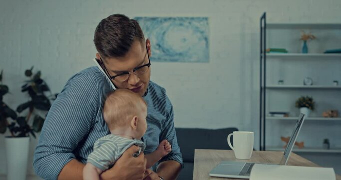handsome father using laptop while talking on smartphone and holding infant