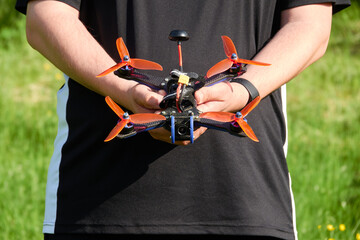 Black drone also race quad, with orange propeller, is held by human hands. Blurred grass in the...