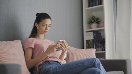 Young charming brunette woman sitting on the gray sofa with pink pillows, listening to the music on the smart phone via white headphones