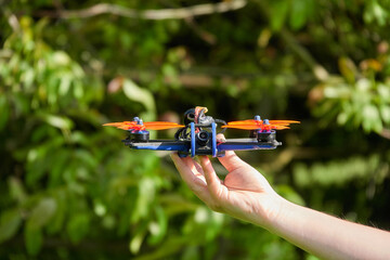 Black drone also race quad, with orange propeller, is held by human hands. Blurred grass in the...