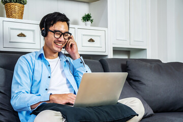 Asian young business man is sitting on the sofa in his own house is talking in a video conference on line with a headset with microphone and laptop in home, work from home