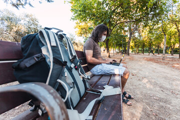 Young man with face mask uses laptop alone while keeping social distance to prevent infectious diseases such as coronavirus in the shade of a park with trees in Spain. Selective focus.