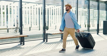 Caucasian young stylish man traveller in hat and with backpack walking at bus station and carrying suitcase on wheels. Handsome male tourist strolling outdoor from train station or aeroport.