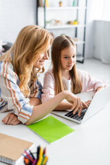 Selective focus of smiling mother using laptop near daughter during webinar at home