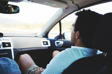 Rear view of young man enjoying comfortable safe trip while sitting on front seat of a luxury car, male passenger monitors the road with automobile during long awaited summer weekend with his friends
