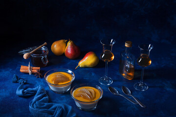 Obraz na płótnie Canvas Panna cotta Italian traditional dessert with pears poached in rum and honey syrup. Dark blue background table, glasses of rum, jar of honey, pears and spice. Dark blue background.