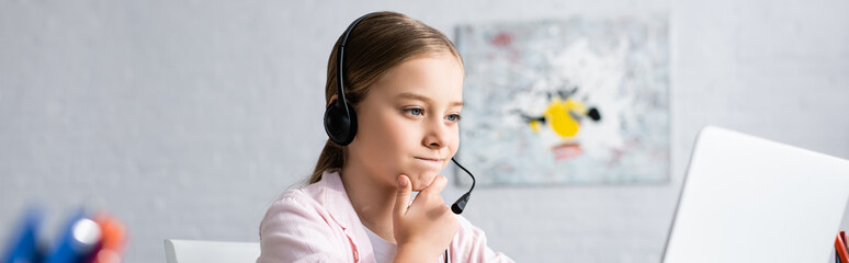 Panoramic crop of pensive kid in headset looking at laptop