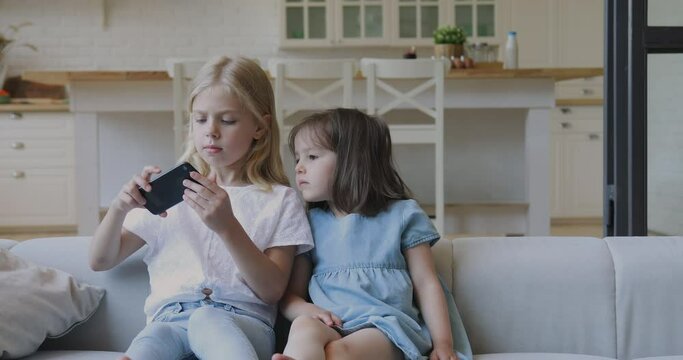 Two funny cute girls sisters learning using smartphone taking selfie at home, little kids siblings holding phone enjoy mobile social media app play game sit on sofa, children and technology concept