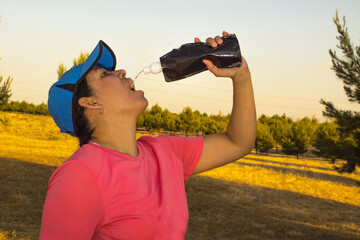 Fototapeta na wymiar Young Caucasian woman wearing sportswear a brushed shirt and a blue cap drinking water from a black bottle after running