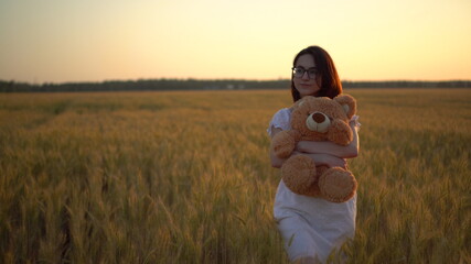 A young woman walks through a wheat field with a teddy bear at sunset. Girl hugs a teddy bear in...
