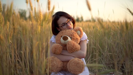 A young woman sits in a dried wheat field with a teddy bear. Girl hugs a teddy bear in hands front...