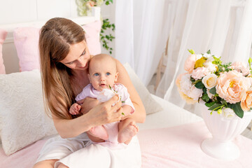 a young beautiful mother with a 6-month-old daughter in her arms sits on a white bed in flowers and hugs her, a place for text