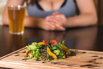 girl sitting at the table in front of her salad of greens, orange, tomato, veal, fresh vegetables on a wooden tray, blackboard, dish. Horizontal photo