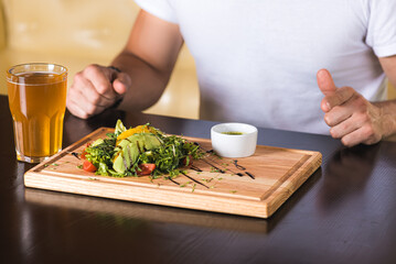 emotional young man sitting at the table, waiting to eat, in front of him a vegetable salad with greens and avocado and meat