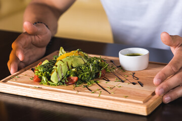 emotional young man sitting at the table, waiting to eat, in front of him a vegetable salad with greens and avocado and meat