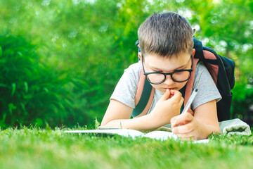 Concentrated schoolboy toddler laying on grass writing in exercise book making homework. male pupil kid drawing writing math outdoors. school education. Back to school. Child in glasses.