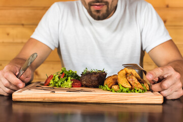 emotional young man sitting at the table, waiting to eat, in front of her is a steak with french fries and a salad of greens