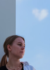 Portrait of a modern single girl.Sad and dreams of meeting love.Day of lovers.Looking off to the side.Wall street,the blue sky and cloud in the shape of a heart.copy space.vertical orientation.