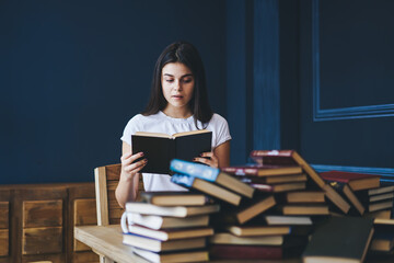 Surprised young woman with brunette hair holding book in hands and reading interesting information sitting at useful literature in university library.Skilled female learning to improve knowledge