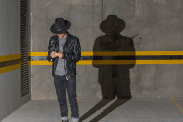 Young latin man with leather jacket, hat and long shadow looking his smartphone in underground car parking