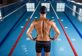 Professional swimmer is wearing a swim cap and swimming shorts, he standing in front of the pool, back view. Start of swimming pool training