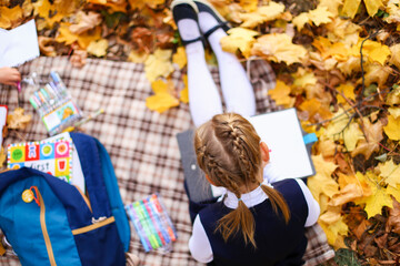 Back to elementary school. Girl in dress does homework with pen in hand.Kid study on plaid blanket.Picnic in park on maple yellow leaves.Blue backpack,notebooks,textbooks lie on ground.Autumn concept