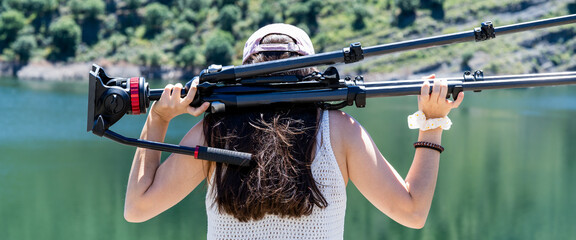 girl carries on her shoulder a professional tripod of photography and video by a lake in summer holidays © JuanFrancisco