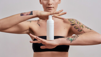 Cropped shot of tattooed woman advertising, holding white plastic bottle with skin care product...
