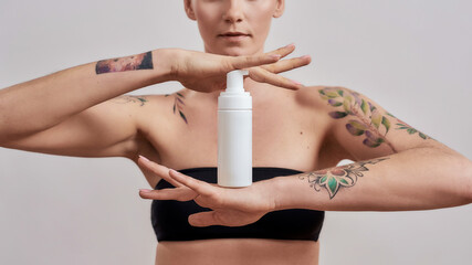 Cropped shot of tattooed woman advertising, holding white plastic bottle with skin care product...
