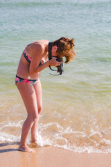 Attractive young woman with dark hair making photo tide on a sandy beach. Outdoor portrait of tanned female photographer working on sandy beach in morning.