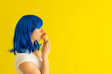 A beautiful woman with blue hair stands in profile and licks a horn of raspberry ice cream on a yellow background. The girl in the wig enjoys eating gelato in a waffle cup. Summer cooling dessert.