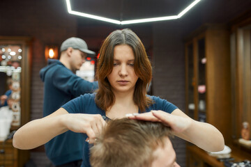 Young professional barber girl making haircut for client sitting in a barbershop chair. Focus on a woman