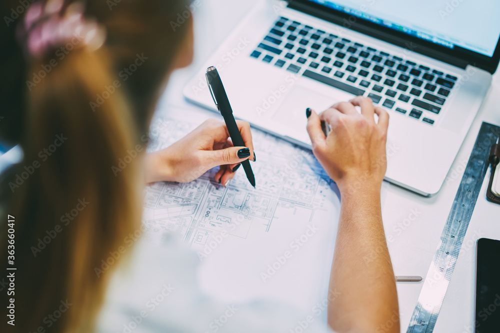 Poster cropped back view of young woman sitting at desktop with building blueprint and modern laptop comput